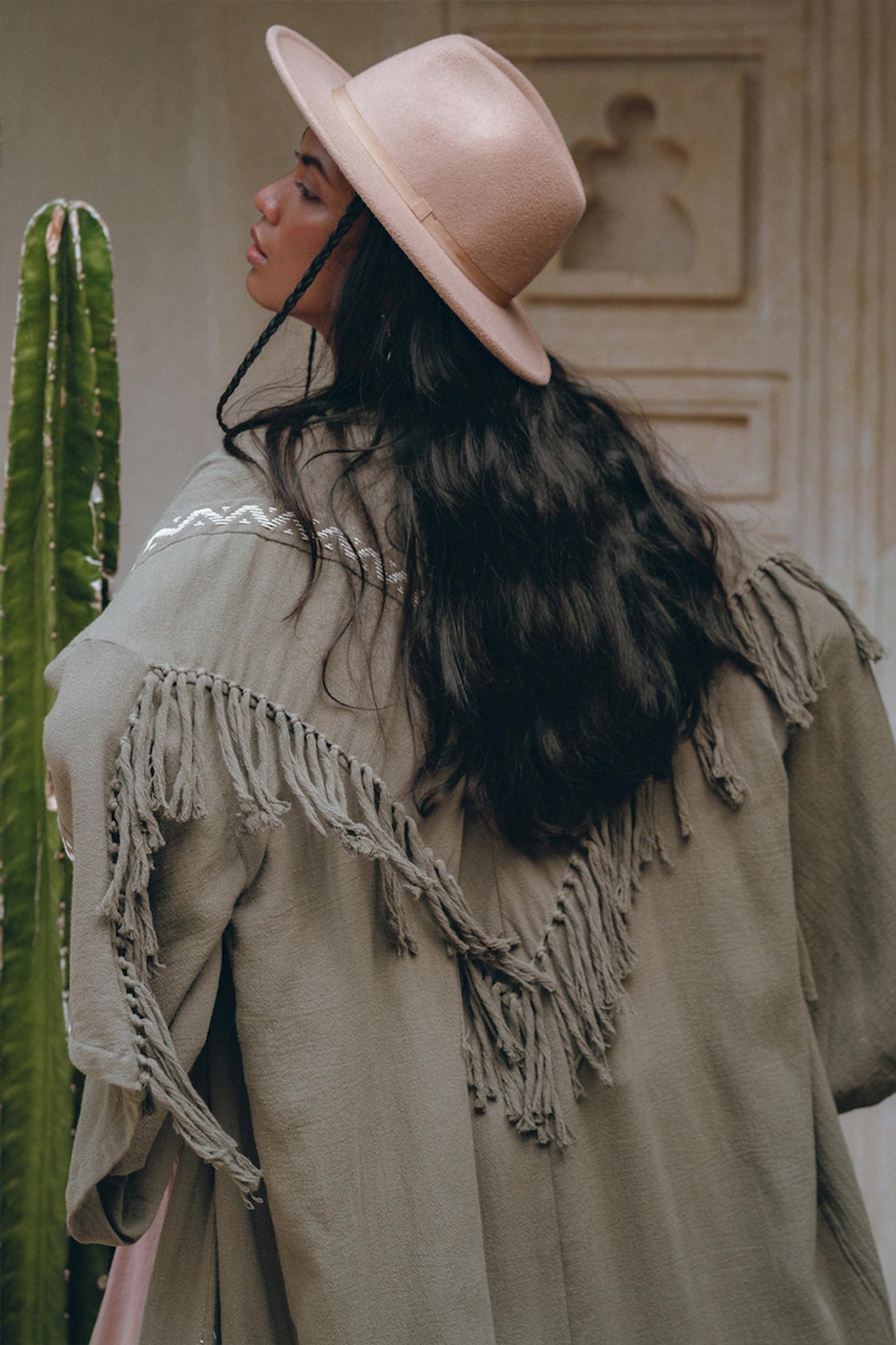 A woman with long dark hair, wearing the AYA Sacred Wear wide-brimmed pink hat and a Sage Colour Organic Cotton Poncho Cape featuring bohemian shamanic embroidery, stands with her back to the camera. A tall cactus is beside her, and a decorative wall serves as the backdrop.