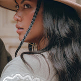 A woman with long, dark hair styled in a single braid looks off to the side, sporting a wide-brimmed beige hat and the Sage Colour Organic Cotton Poncho Cape by AYA Sacred Wear, featuring shamanic embroidery patterns. The background is gently blurred.
