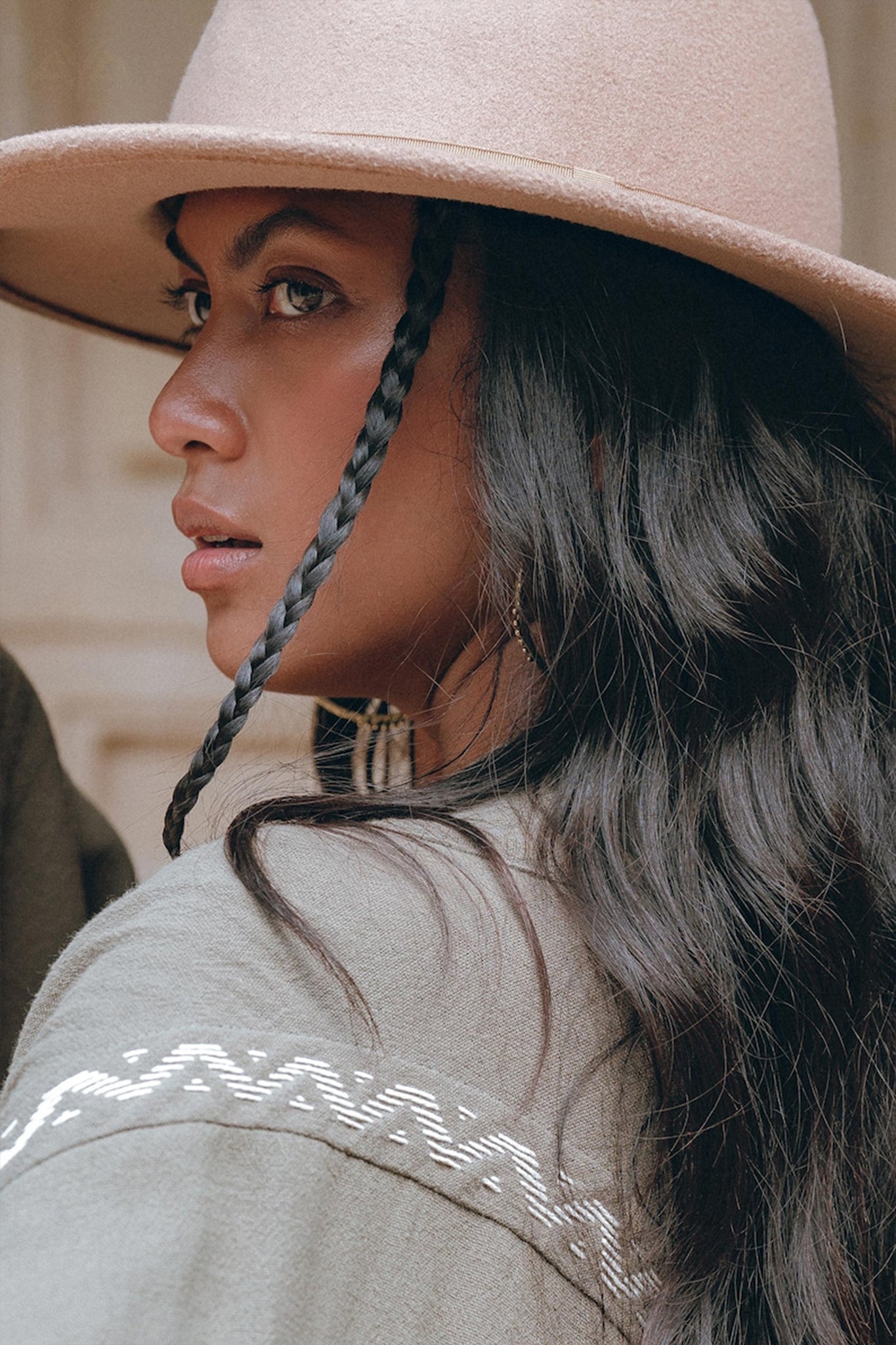 A woman with long, dark hair styled in a single braid looks off to the side, sporting a wide-brimmed beige hat and the Sage Colour Organic Cotton Poncho Cape by AYA Sacred Wear, featuring shamanic embroidery patterns. The background is gently blurred.