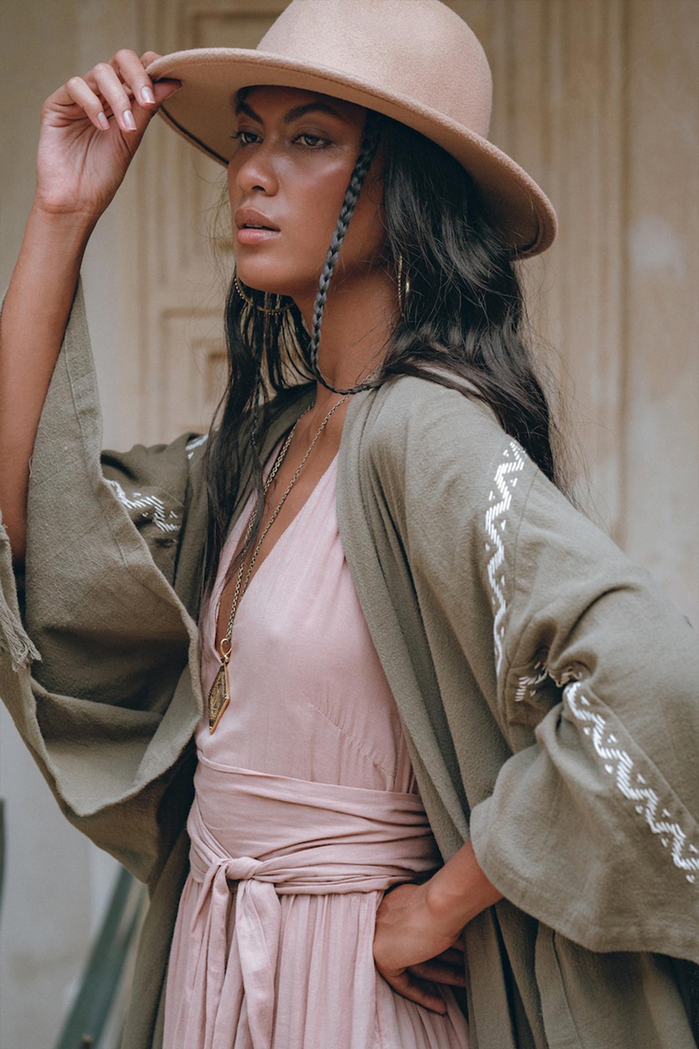 A person wearing a beige hat and an AYA Sacred Wear Sage Colour Organic Cotton Poncho Cape draped over a light pink dress stands in a relaxed pose. Their long dark hair complements the layered necklaces, while the background is softly blurred, emphasizing their serene presence.