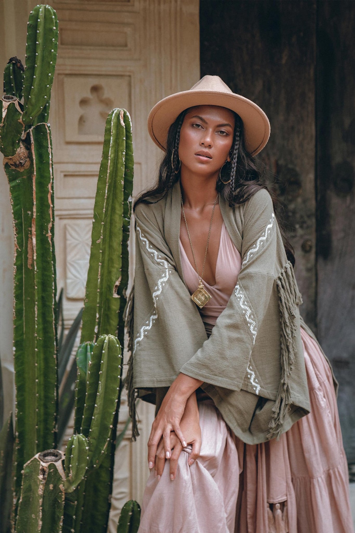 A woman in a chic ensemble leans against a tall cactus, donning a wide-brimmed hat and the Sage Colour Organic Cotton Poncho Cape by AYA Sacred Wear—a green jacket with fringes and shamanic embroidery over a pink dress. In the backdrop, an ornate wooden door with decorative patterns completes the scene.