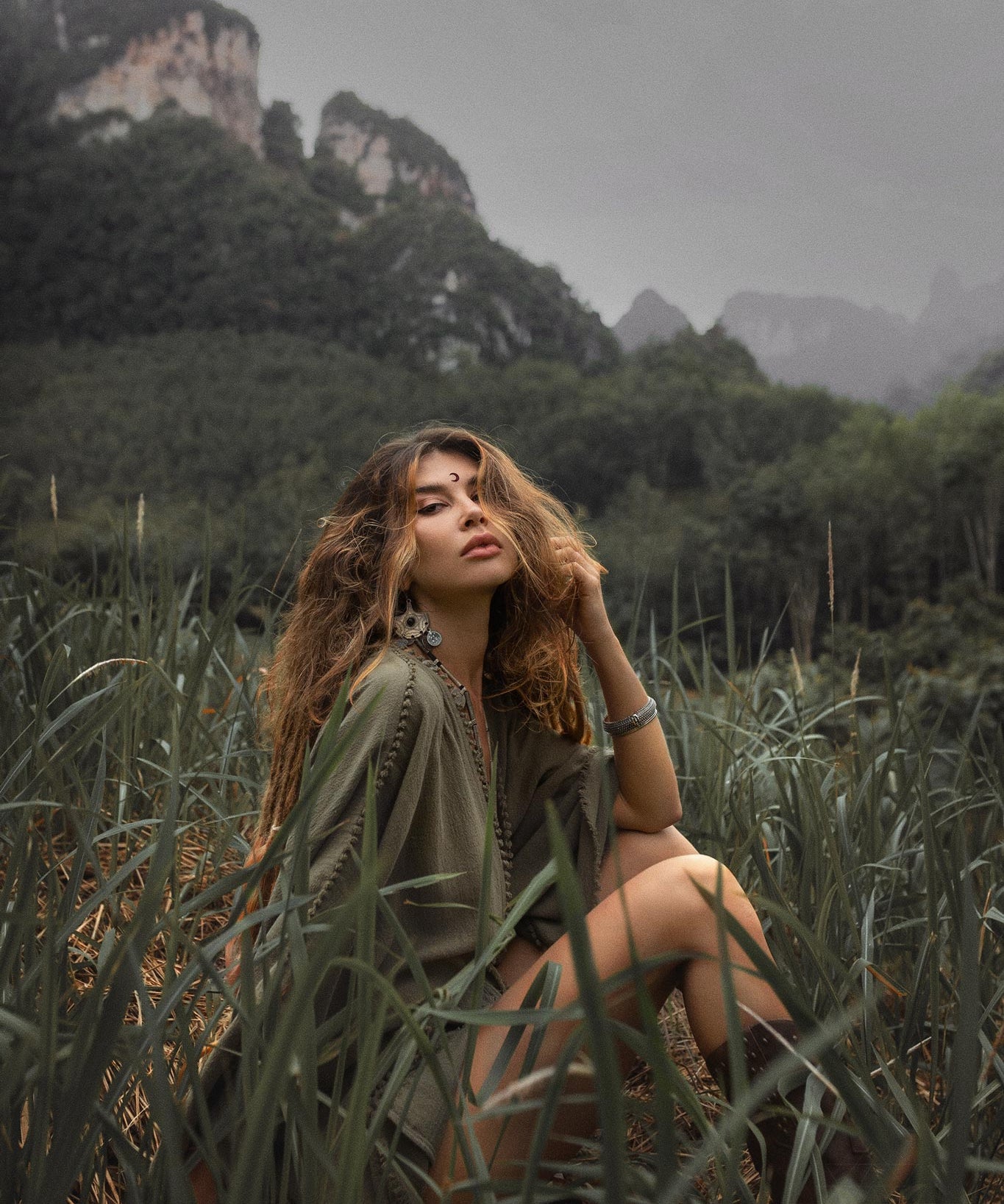 A woman with wavy hair in a sage green, hand-knotted romper sits in tall grass with a mountainous landscape in the background.