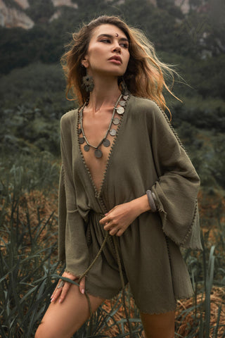 A woman standing outdoors, wearing a sage green hand-knotted mini jumpsuit romper dress. The organic fabric drapes loosely, with a deep V-neckline and bell sleeves. She accessorizes with large statement earrings, a necklace with round medallions, and a bracelet. Her wavy hair flows in the wind as she gazes into the distance, surrounded by lush greenery in a natural, forest-like setting.