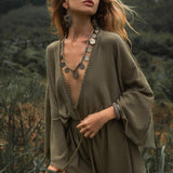 A woman standing outdoors, wearing a sage green hand-knotted mini jumpsuit romper dress. The organic fabric drapes loosely, with a deep V-neckline and bell sleeves. She accessorizes with large statement earrings, a necklace with round medallions, and a bracelet. Her wavy hair flows in the wind as she gazes into the distance, surrounded by lush greenery in a natural, forest-like setting.