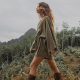 A woman walking through a natural outdoor environment, wearing a sage green hand-knotted mini organic jumpsuit romper dress. The outfit features flowing bell sleeves and a cinched waist. She completes her look with tall brown boots adorned with studs. Her long, wavy hair flows naturally as she strides confidently across the earthy terrain, with a backdrop of green trees and distant hills under a cloudy sky.