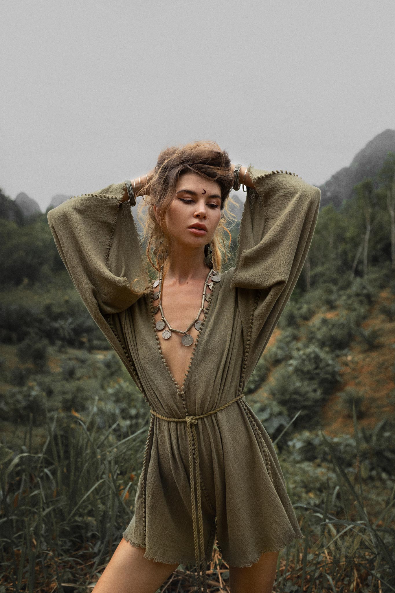 A woman standing outdoors with her hands raised behind her head, wearing a sage green hand-knotted mini organic jumpsuit romper dress. The outfit has wide bell sleeves and a cinched waist. She accessorizes with a necklace featuring round medallions and large earrings. Her expression is serene, with her hair styled in a loose updo, and she is surrounded by lush greenery with distant hills and overcast skies in the background.