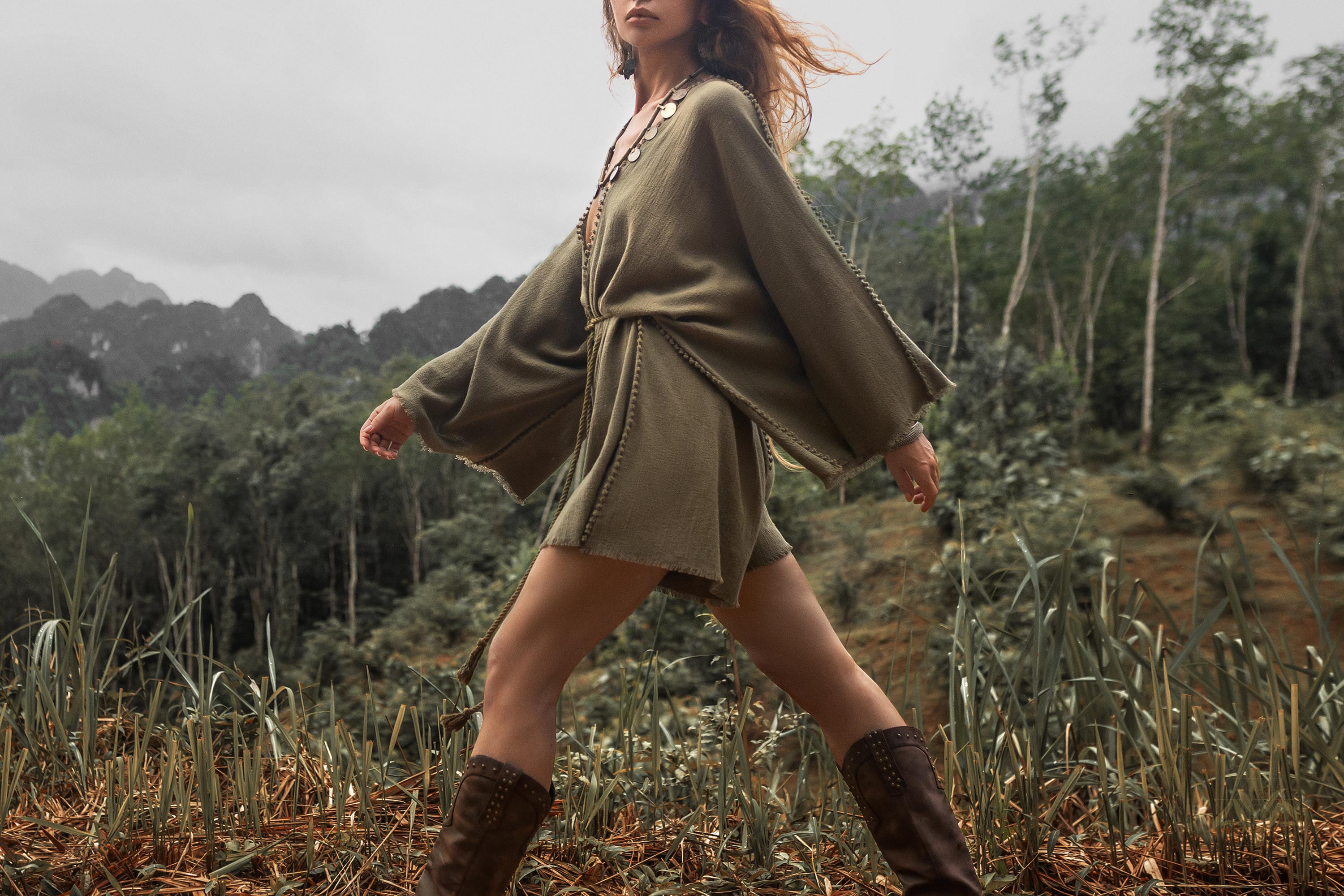 A person walks through a lush, mountainous landscape wearing a sage green hand-knotted jumpsuit from AYA Sacred Wear and brown boots. The sky is overcast, and the background features dense greenery and tall, rocky mountains. The individual's hair flows freely as they move in a comfortable summer style.