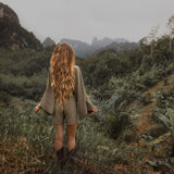 A person with long, wavy hair stands with their back to the camera, wearing a comfortable summer style Sage Green Hand Knotted Jumpsuit from AYA Sacred Wear and boots. They are in a lush, green landscape with dense foliage, grasses, and distant mist-covered mountains. The sky is overcast, adding a dreamy atmosphere.