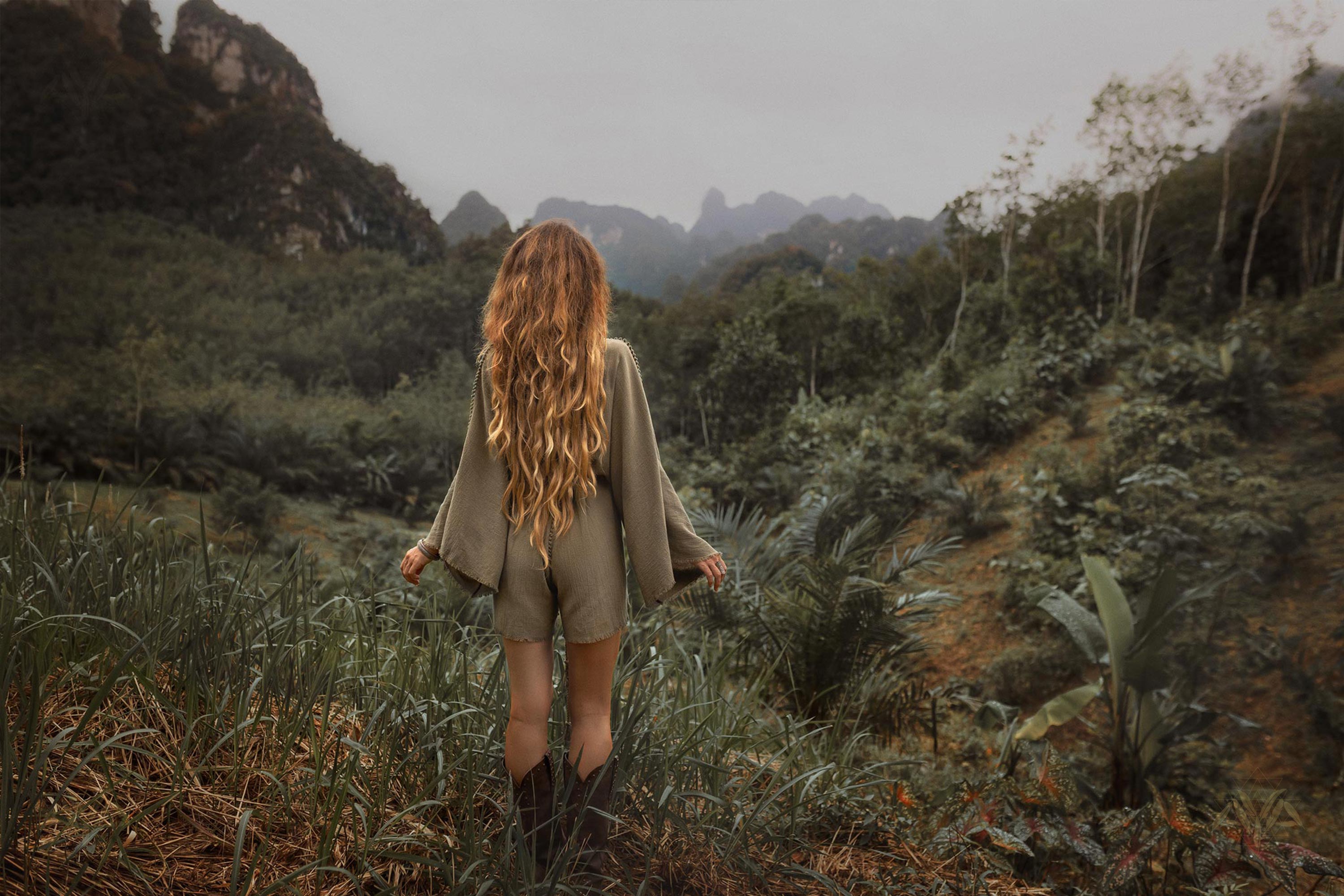 A person with long, wavy hair stands with their back to the camera, wearing a comfortable summer style Sage Green Hand Knotted Jumpsuit from AYA Sacred Wear and boots. They are in a lush, green landscape with dense foliage, grasses, and distant mist-covered mountains. The sky is overcast, adding a dreamy atmosphere.
