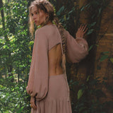 A woman with long, wavy, braided hair stands in a lush forest wearing the AYA Sacred Wear Samudra Dress in Blush Rose—a backless boho maxi. She glances over her shoulder, hand on a vine-draped tree trunk, brass spiral bracelet on her wrist.