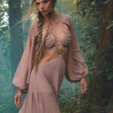 A woman with long, braided hair stands in a misty forest, wearing the AYA Sacred Wear Samudra Dress in Blush Rose—a backless boho maxi with gathered sleeves and cutout top—paired with a handmade brass spiral pendant.