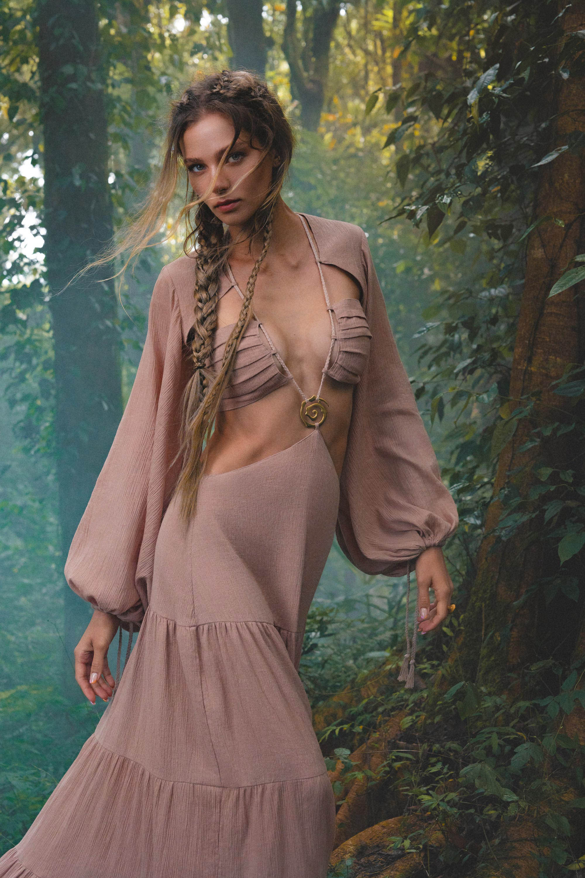 A woman with long, braided hair stands in a misty forest, wearing the AYA Sacred Wear Samudra Dress in Blush Rose—a backless boho maxi with gathered sleeves and cutout top—paired with a handmade brass spiral pendant.