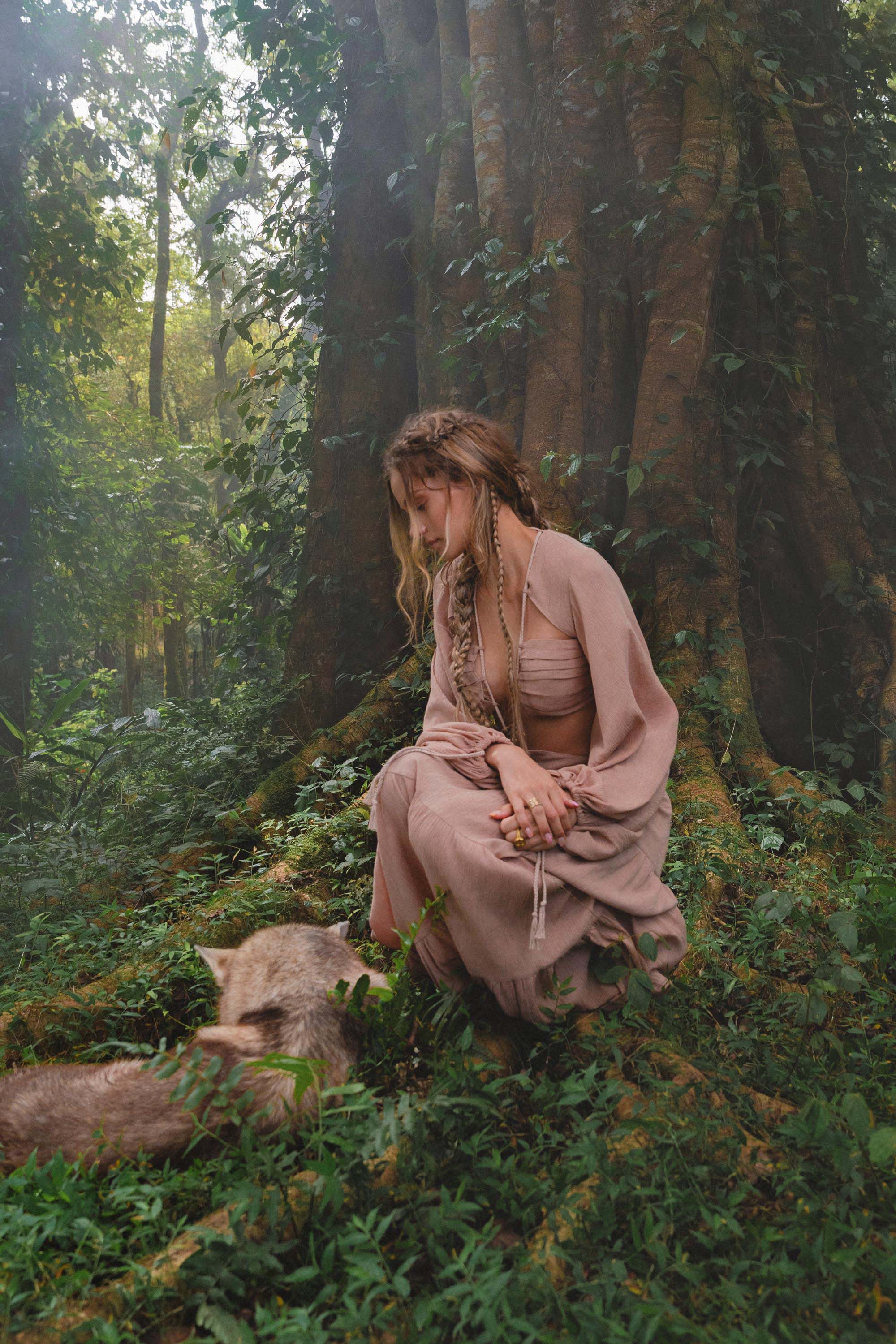 A woman in a blush rose Samudra Dress by AYA Sacred Wear kneels on mossy forest ground, her braided hair adorned with leaves and brass spiral, gently facing a raccoon amid lush foliage and a large tree trunk.