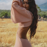 A woman with long wavy hair poses in a dry, grassy field wearing the AYA Sacred Wear Samudra Dress in Blush Rose—a backless boho maxi dress—paired with a handmade brass spiral. Green trees and distant mountains frame the scene.