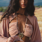 A woman with long, wavy dark hair stands outdoors in a blush rose Samudra Dress by AYA Sacred Wear, featuring voluminous sleeves and a gathered bodice. She accessorizes with a handmade brass spiral pendant on a rope necklace, looking serene and confident.