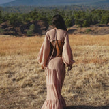 A woman with loose dark hair in an AYA Sacred Wear Samudra Dress in Blush Rose, a backless boho maxi, walks away through dry grass toward distant mountains under a partly cloudy sky, green trees at the base.