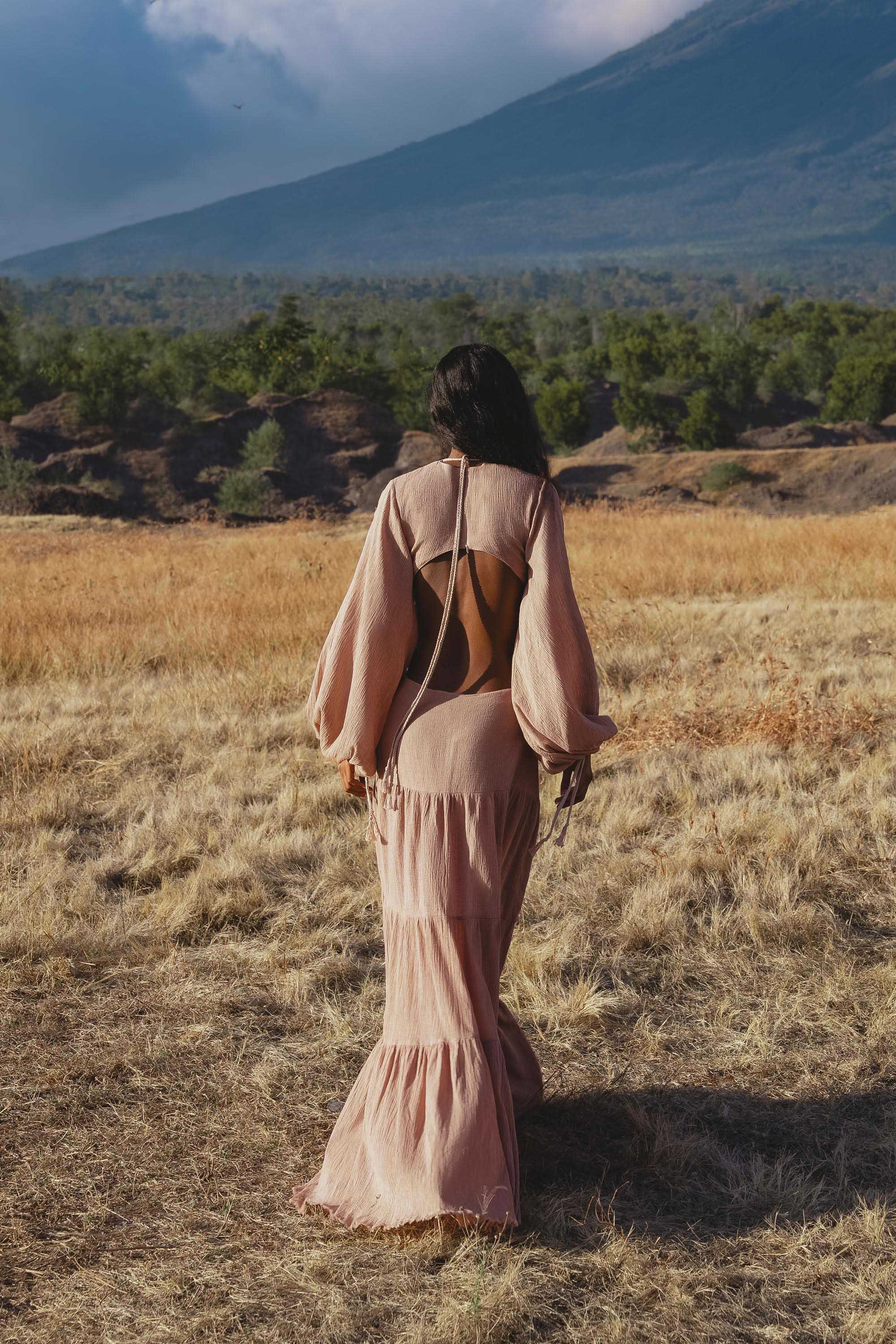 A woman with loose dark hair in an AYA Sacred Wear Samudra Dress in Blush Rose, a backless boho maxi, walks away through dry grass toward distant mountains under a partly cloudy sky, green trees at the base.