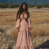 A woman with long, wavy hair stands in a dry grassy field wearing the AYA Sacred Wear Samudra Dress in Blush Rose—a backless boho maxi dress. Mountains and trees rise in the background beneath a partly cloudy sky.