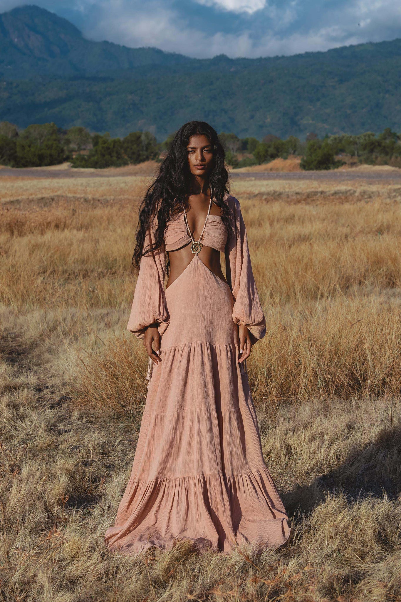 A woman with long, wavy hair stands in a dry grassy field wearing the AYA Sacred Wear Samudra Dress in Blush Rose—a backless boho maxi dress. Mountains and trees rise in the background beneath a partly cloudy sky.