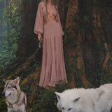 A woman in an AYA Sacred Wear Samudra Dress in Blush Rose, a backless boho maxi with a deep neckline, stands by a tree in a lush forest. At her feet are white and gray wolves, and her look is finished with a handmade brass spiral.