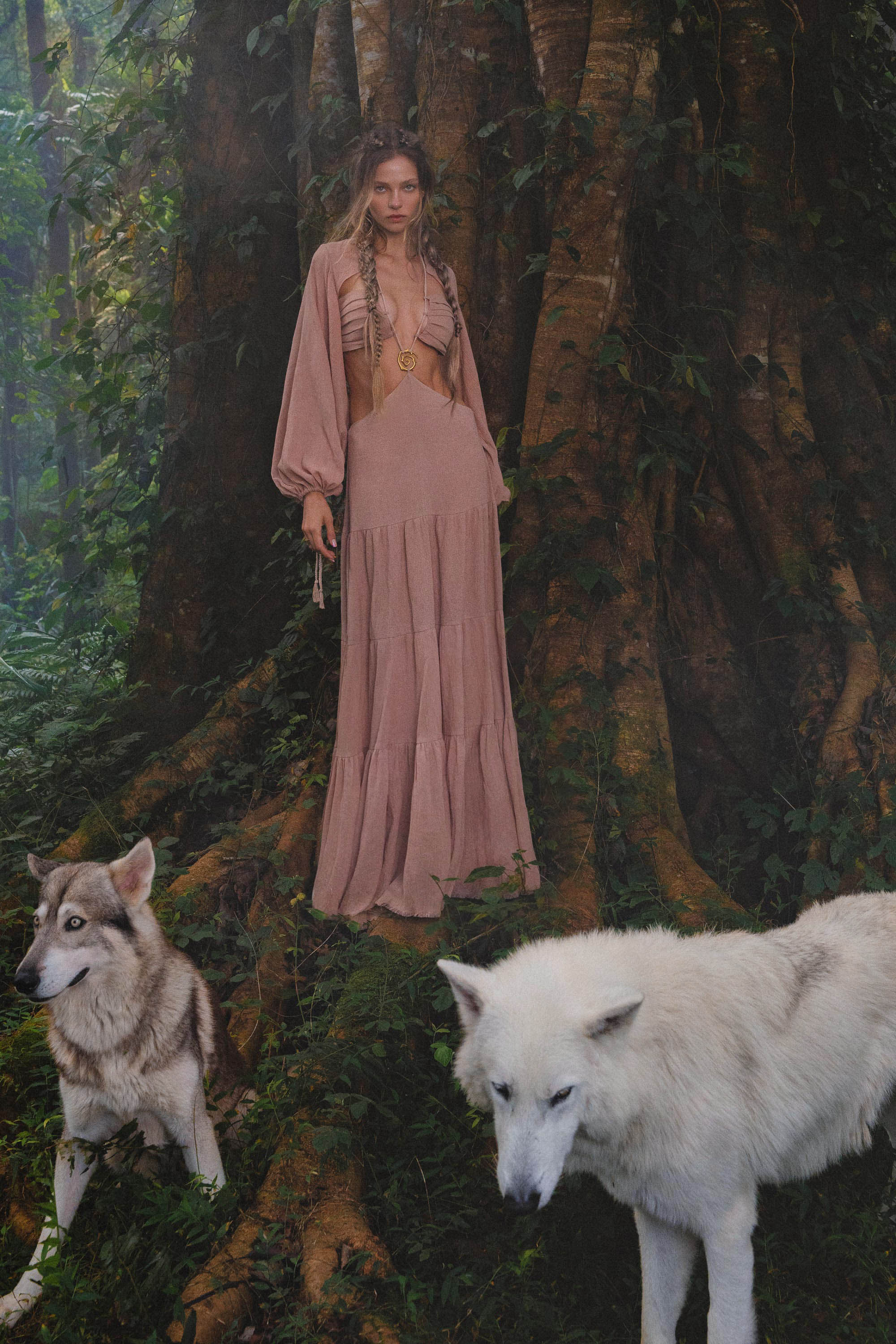 A woman in an AYA Sacred Wear Samudra Dress in Blush Rose, a backless boho maxi with a deep neckline, stands by a tree in a lush forest. At her feet are white and gray wolves, and her look is finished with a handmade brass spiral.