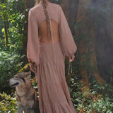A woman with long braided hair stands in a lush forest, wearing the flowing Samudra Dress in Blush Rose by AYA Sacred Wear. Beside her, a wolf-like dog faces away, both framed by greenery and a large tree trunk.