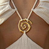 A close-up shows a tan individual wearing the AYA Sacred Wear Samudra Dress in Ivory, an organic bohemian linen-cotton dress with thin straps. A handmade brass spiral pendant on braided white cords rests above the open back.
