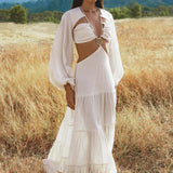 A woman with straight dark hair, calm expression, stands in a dry grass field wearing the Samudra Dress in Ivory Colour by AYA Sacred Wear—an organic bohemian linen-cotton dress with long sleeves, side cut-outs, and brass spiral chest detail. Green hills and mountains behind.
