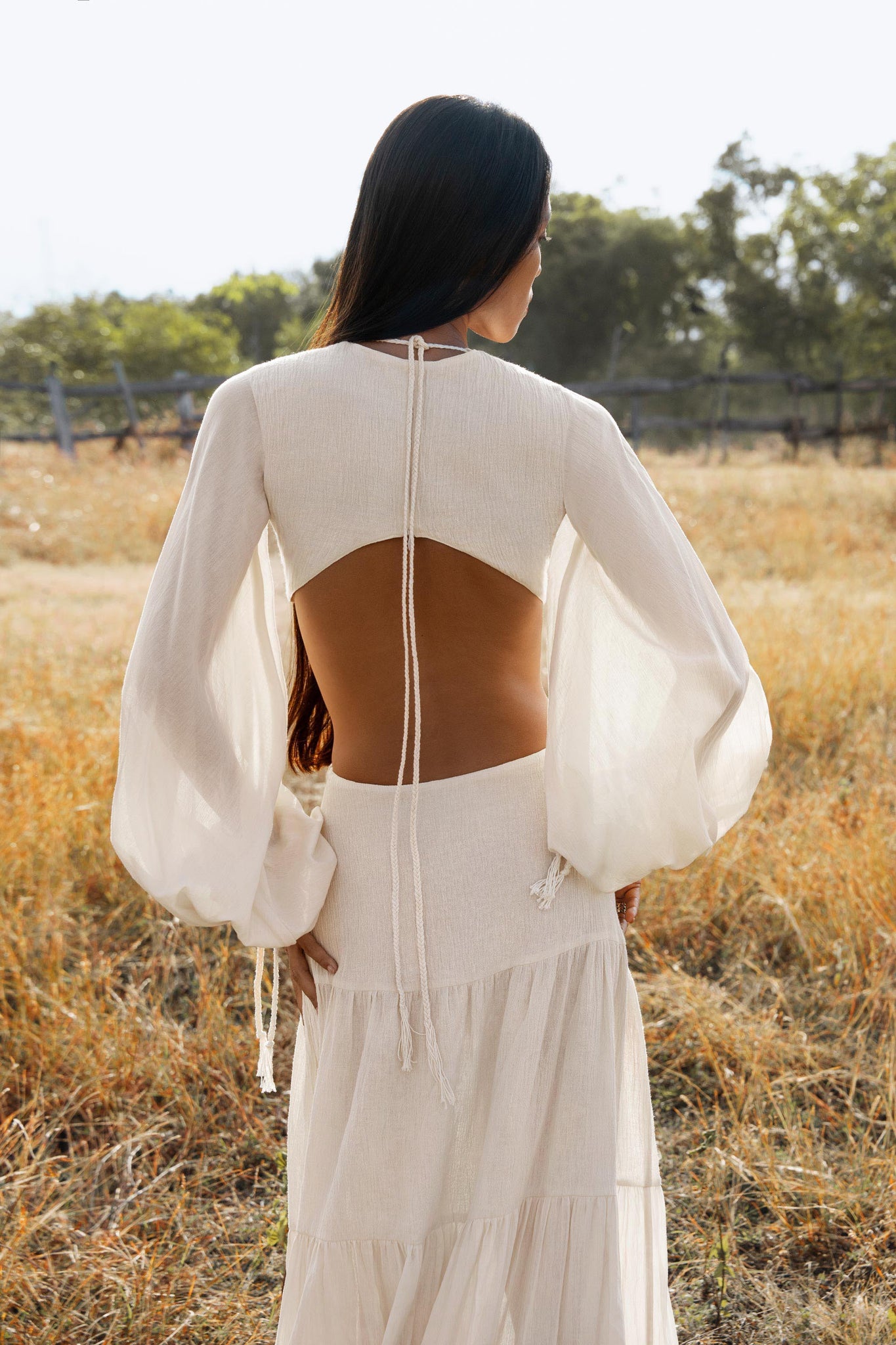 A woman stands in a sunny field, wearing the Samudra Dress in Ivory by AYA Sacred Wear—an organic bohemian dress with sheer sleeves, an open back tied with thin strings, and elegant linen cotton fabric. Her dark hair flows as the background blurs softly.