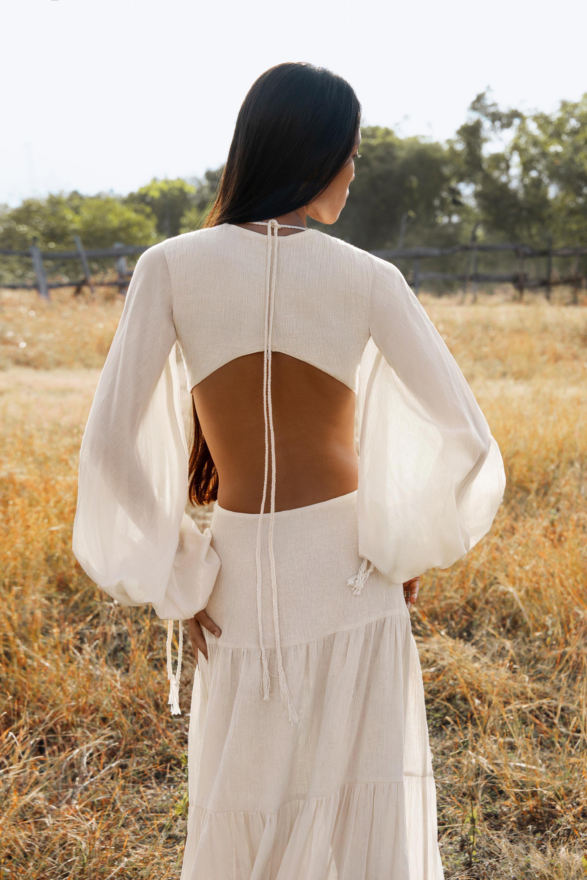 A woman stands in a sunny field, wearing the Samudra Dress in Ivory by AYA Sacred Wear—an organic bohemian dress with sheer sleeves, an open back tied with thin strings, and elegant linen cotton fabric. Her dark hair flows as the background blurs softly.