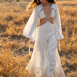 A woman with long, wavy dark hair walks through a sunlit field in the Samudra Dress in Ivory Colour by AYA Sacred Wear–an organic bohemian dress with a halter top and tiered skirt. The soft light highlights her serene expression amidst the earthy landscape.
