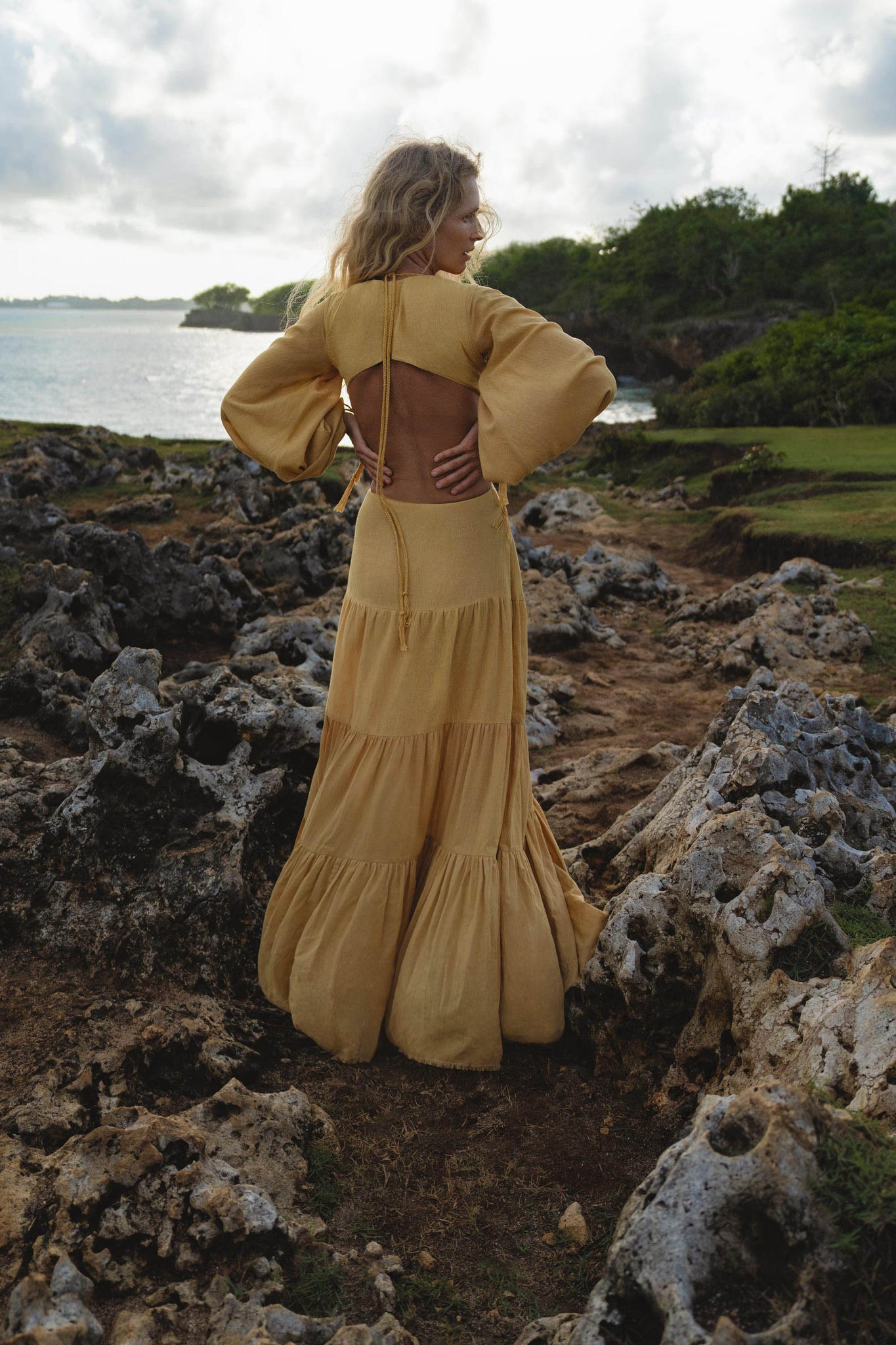 A woman with wavy blonde hair stands on rocky seaside terrain, facing away. She wears the AYA Sacred Wear Samudra Dress in Saffron Gold Colour – an organic, backless boho maxi dress with loose sleeves. Lush greenery surrounds her under a cloudy sky.