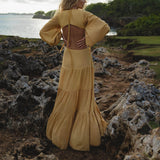 A woman with wavy blonde hair stands on rocky seaside terrain, facing away. She wears the AYA Sacred Wear Samudra Dress in Saffron Gold Colour – an organic, backless boho maxi dress with loose sleeves. Lush greenery surrounds her under a cloudy sky.