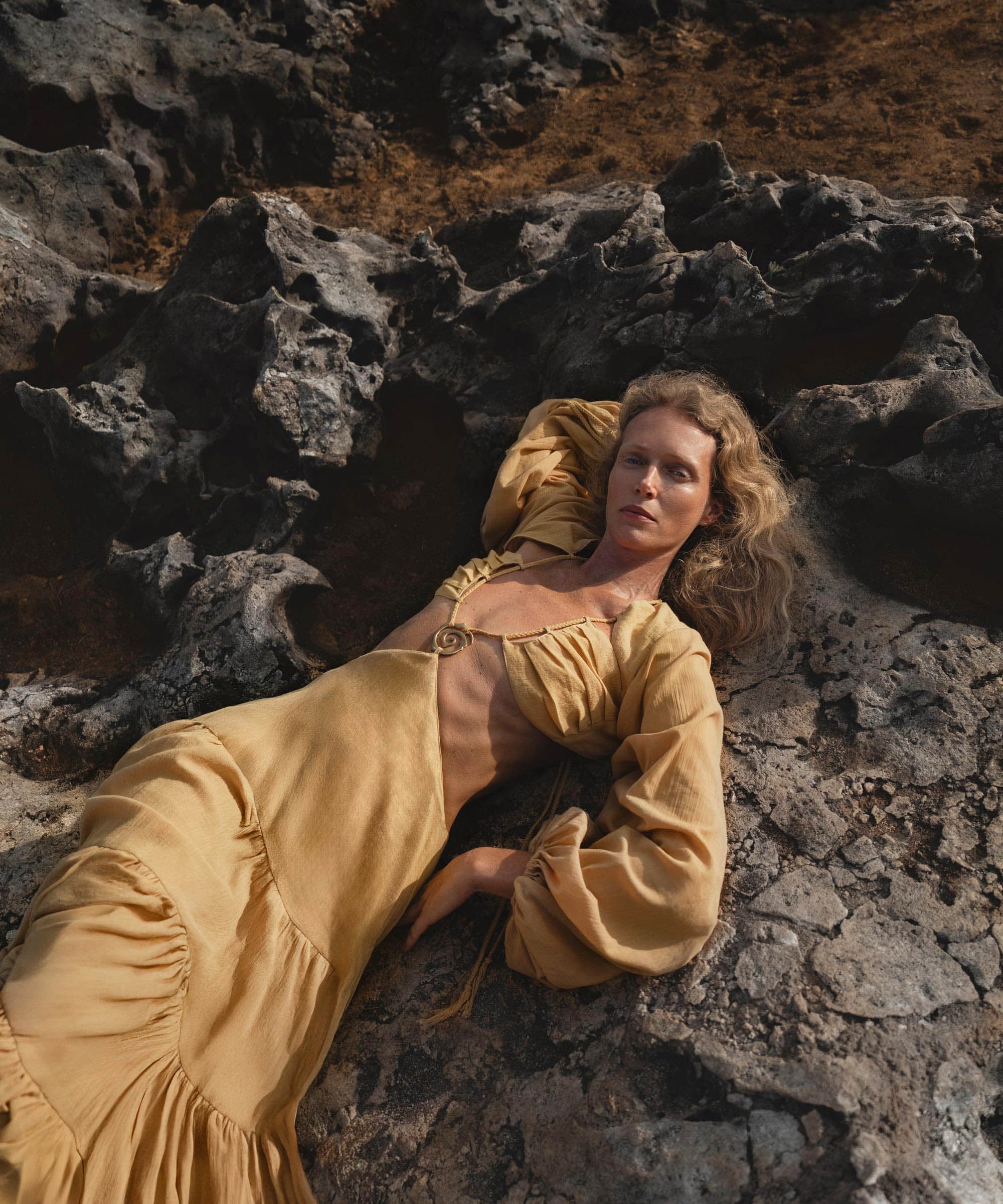 A woman with wavy blonde hair reclines on dark volcanic rocks, wearing AYA Sacred Wear’s Samudra Dress in Saffron Gold—a flowing organic backless boho maxi with a deep neckline and gathered waist—her direct gaze contrasting the rugged earth.