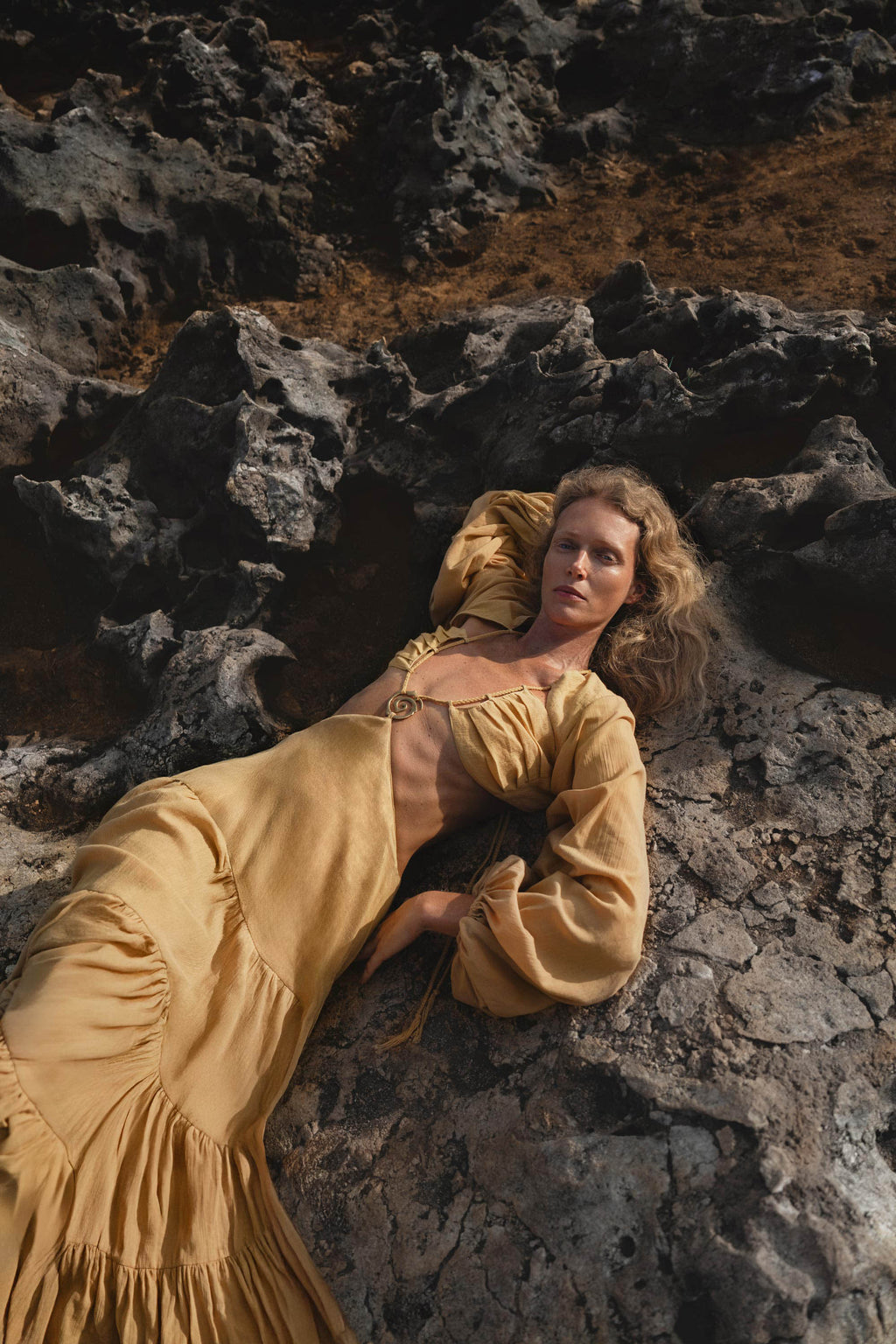 A woman with wavy blonde hair reclines on dark volcanic rocks, wearing AYA Sacred Wear’s Samudra Dress in Saffron Gold—a flowing organic backless boho maxi with a deep neckline and gathered waist—her direct gaze contrasting the rugged earth.