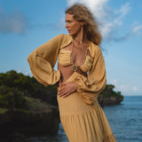 A woman with wavy blonde hair poses outdoors by the sea in AYA Sacred Wear's Samudra Dress in Saffron Gold—a backless, organic boho maxi with billowy sleeves and a long skirt. Rocky cliffs, greenery, and blue sky form the scenic backdrop.