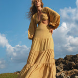 A woman with long wavy blonde hair poses barefoot outdoors in the AYA Sacred Wear Samudra Dress in Saffron Gold – an organic, backless boho maxi dress with long sleeves. The blue sky and scattered clouds complete the scene.