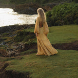 A woman with long blonde hair wears the AYA Sacred Wear Samudra Dress in Saffron Gold—an organic, backless boho maxi—while standing on a grassy cliffside at sunset, with lush greenery and glowing ocean as her backdrop.