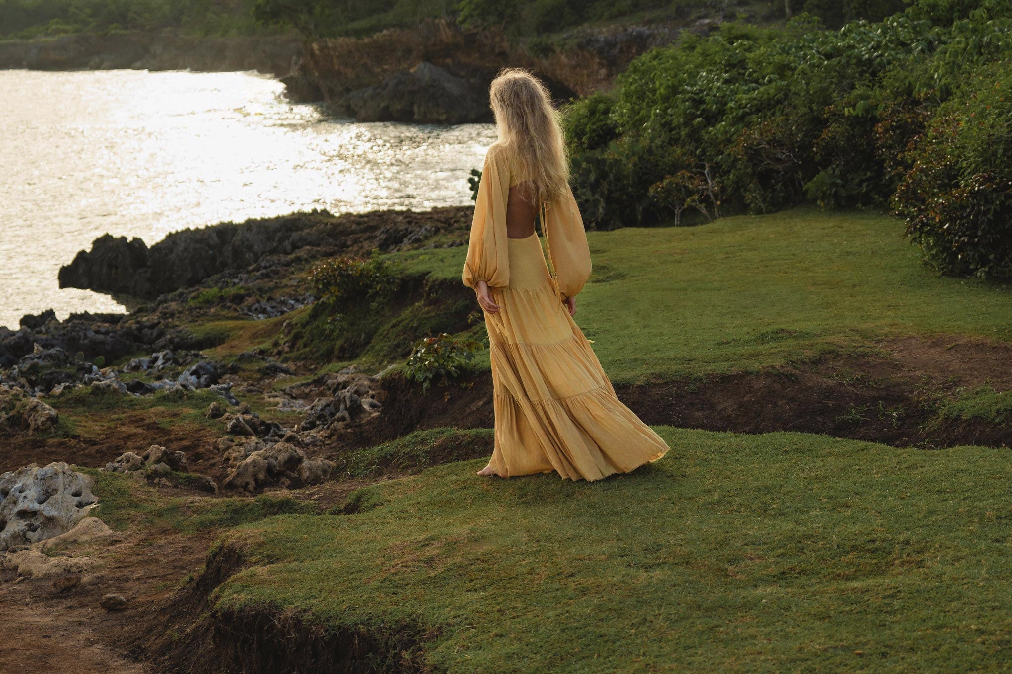 A woman with long blonde hair wears the AYA Sacred Wear Samudra Dress in Saffron Gold—an organic, backless boho maxi—while standing on a grassy cliffside at sunset, with lush greenery and glowing ocean as her backdrop.