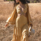 A woman stands in a sunny, grassy field wearing the AYA Sacred Wear Samudra Dress in Saffron Gold, an organic backless boho maxi with long sleeves and cutout bodice. She holds a white wide-brim hat as trees and hills frame the scene.