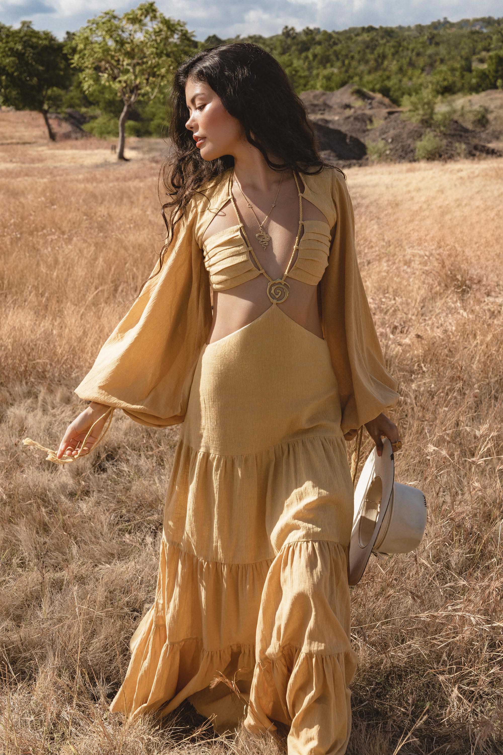 A woman stands in a sunny, grassy field wearing the AYA Sacred Wear Samudra Dress in Saffron Gold, an organic backless boho maxi with long sleeves and cutout bodice. She holds a white wide-brim hat as trees and hills frame the scene.