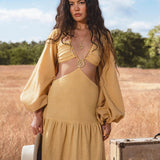 A woman with long, wavy hair wears the Samudra Dress in Saffron Gold by AYA Sacred Wear—an organic backless boho maxi dress with puffed sleeves. She stands in a sunlit field holding a hat by a vintage suitcase, trees and blue sky behind her.