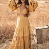 A woman with long wavy hair wears the AYA Sacred Wear Samudra Dress in saffron gold, an organic backless boho maxi with tiered skirt and puff sleeves. She holds a wide-brim hat in a sunlit field as a vintage suitcase rests nearby.