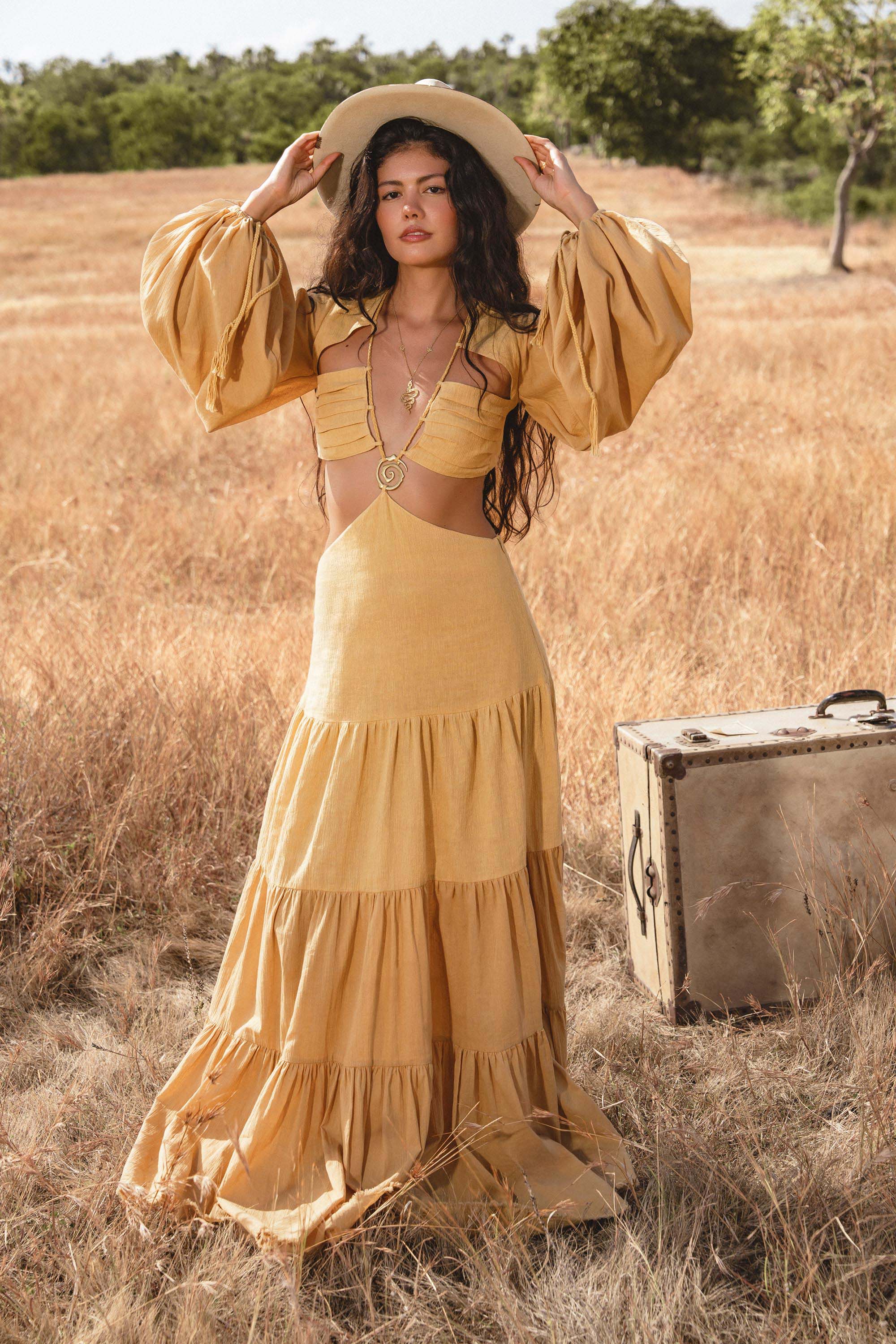 A woman with long wavy hair wears the AYA Sacred Wear Samudra Dress in saffron gold, an organic backless boho maxi with tiered skirt and puff sleeves. She holds a wide-brim hat in a sunlit field as a vintage suitcase rests nearby.