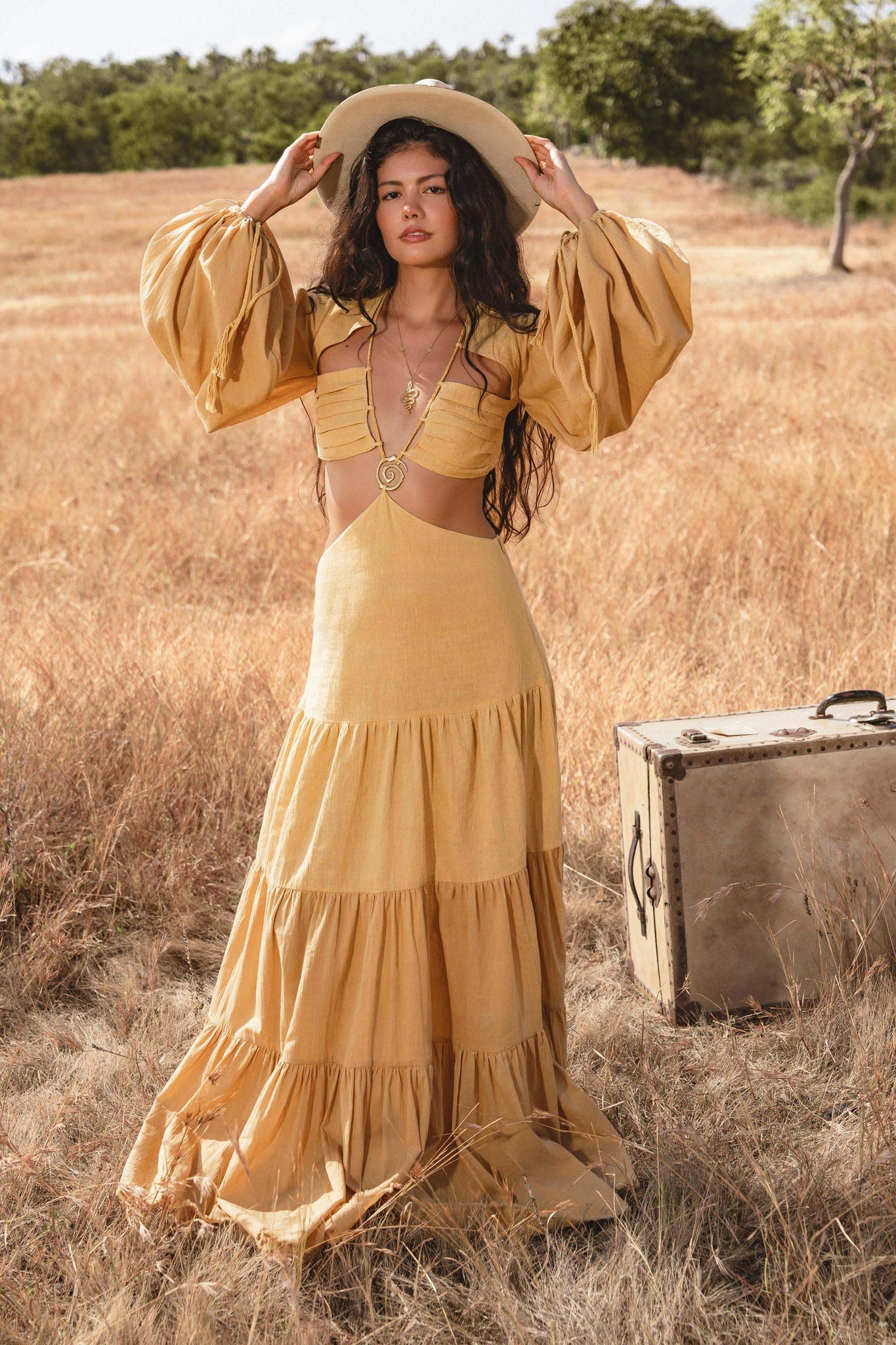 A woman with long wavy hair wears the AYA Sacred Wear Samudra Dress in saffron gold, an organic backless boho maxi with tiered skirt and puff sleeves. She holds a wide-brim hat in a sunlit field as a vintage suitcase rests nearby.