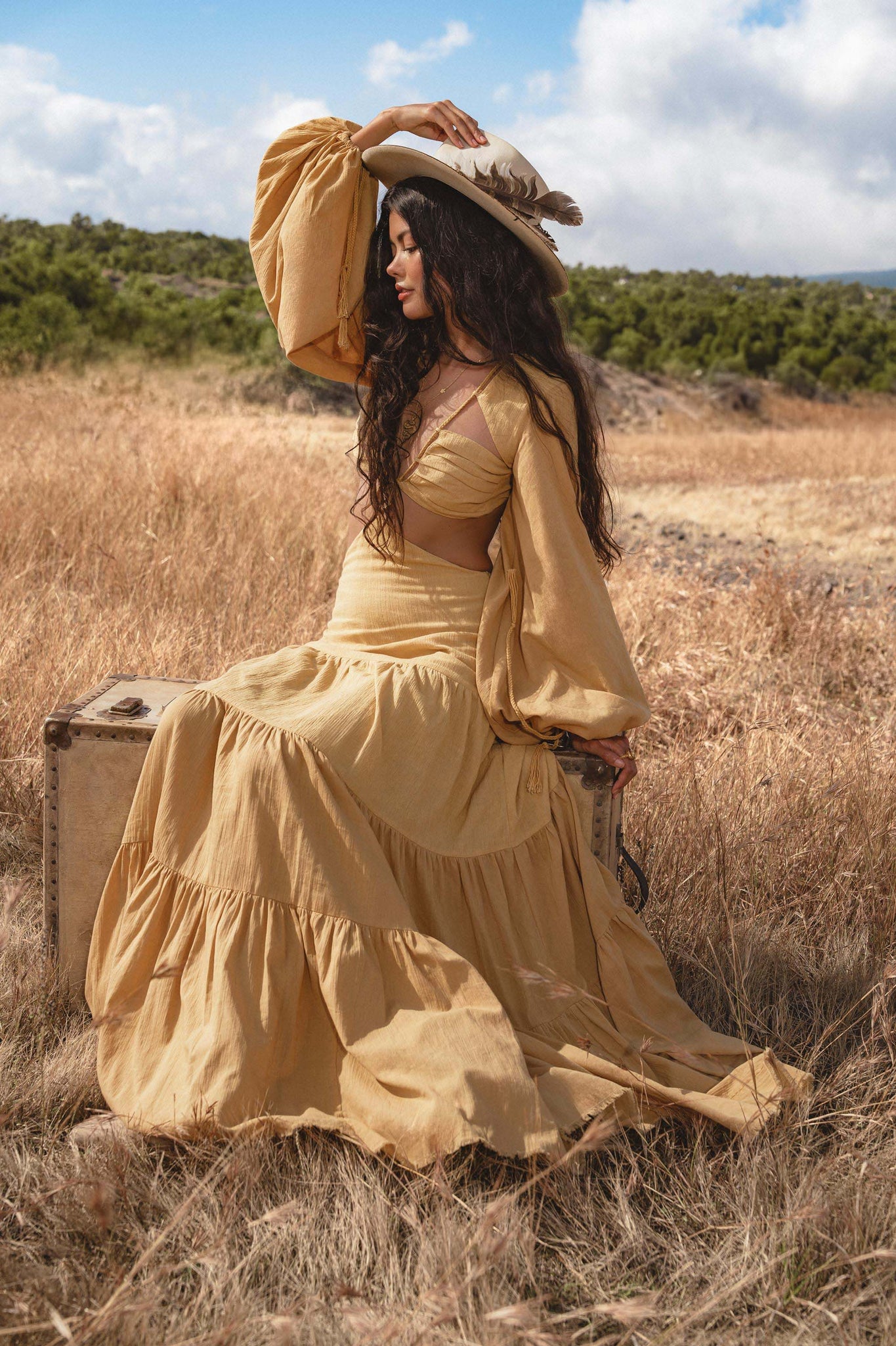 A woman wearing the AYA Sacred Wear Samudra Dress in saffron gold—a flowing, organic, backless boho maxi—sits on a vintage suitcase in a sunlit field, gazing sideways with green trees and blue sky behind her.