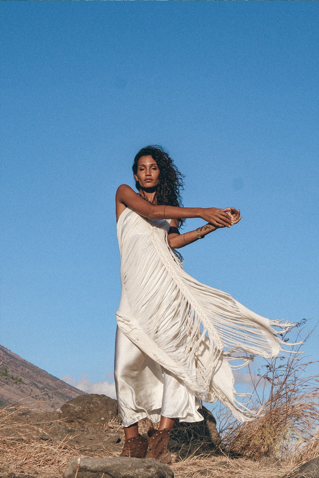 A woman poses against a clear blue sky, wearing the See Through Dress • Long Maxi Dress • Tribal Raw Cotton Cover-up by AYA Sacred Wear. She moves gracefully, with one arm raised and the other extended, amid dry grass and rocks.