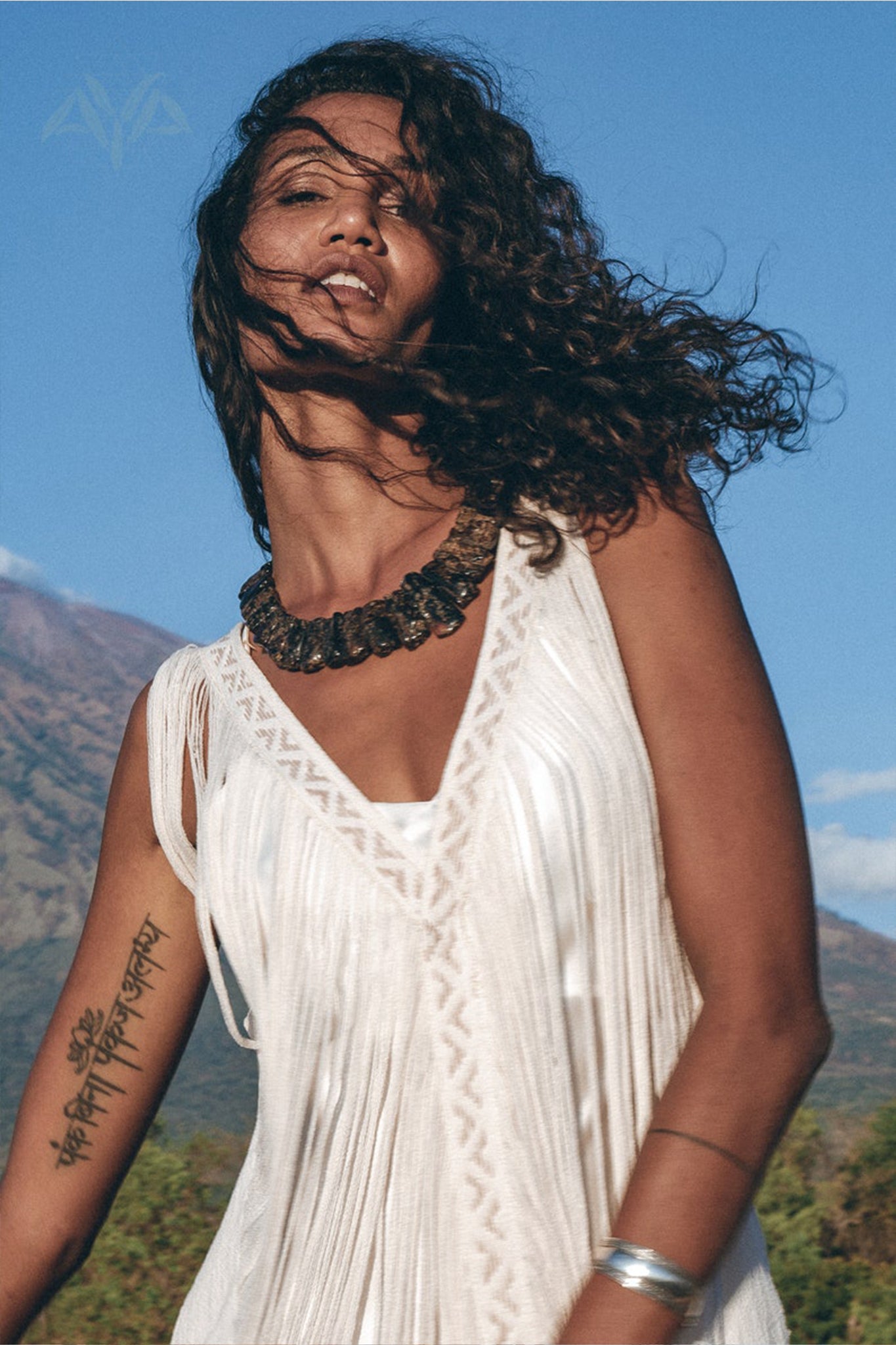 A woman with curly hair poses outdoors in a See Through Dress by AYA Sacred Wear, a long maxi dress designed as a tribal raw cotton cover-up. The mountain and blue sky form the backdrop as her tattooed arm, adorned with a bangle and a statement necklace, completes the look.