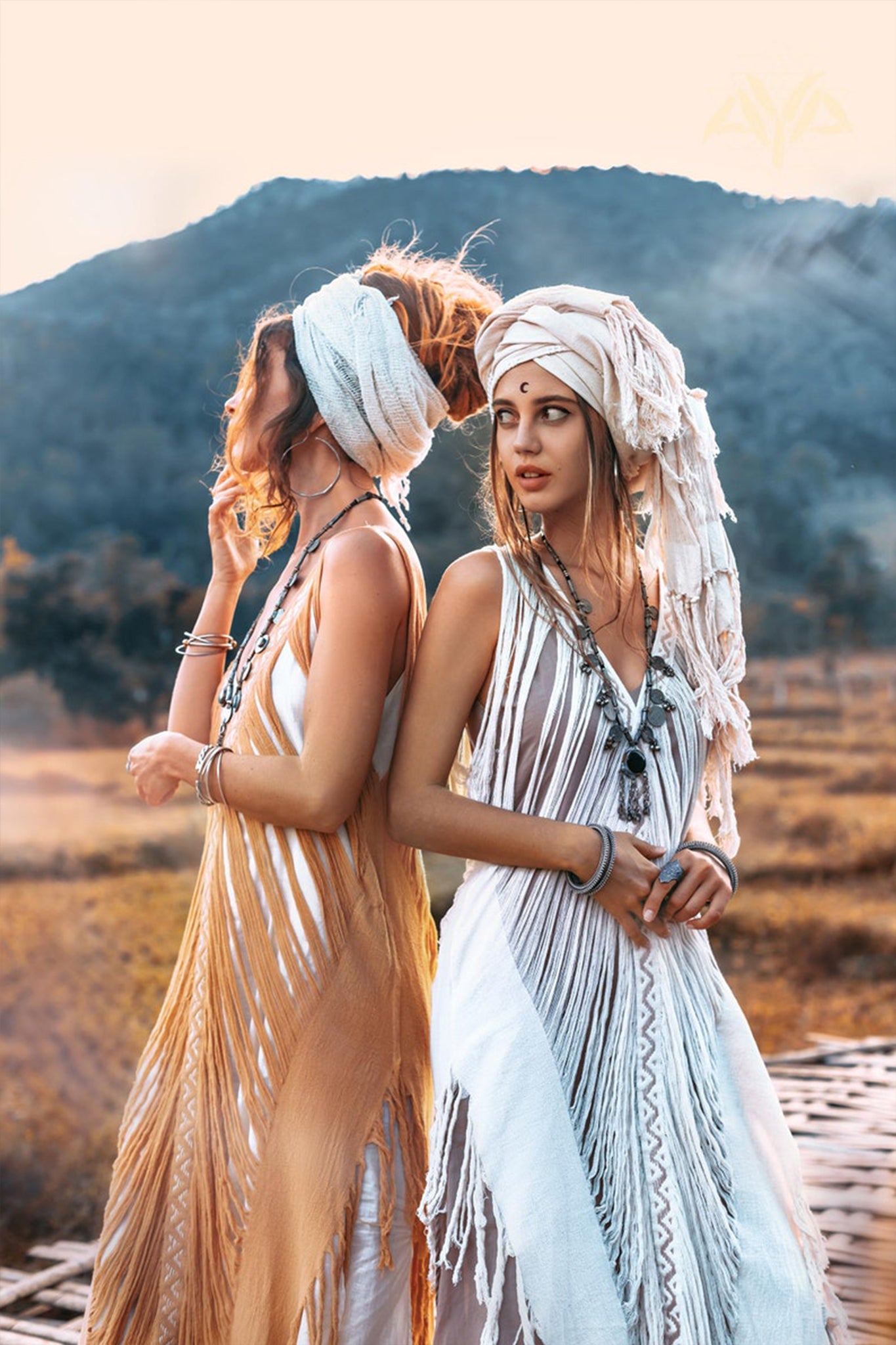 Two women wearing AYA Sacred Wear's Tribal Raw Cotton Cover-ups stand back to back in a field at sunset. One faces left, the other right, with majestic mountains as their backdrop. Their bohemian style is accentuated by layered necklaces and bracelets, complemented by the handmade embroidery of their See Through Long Maxi Dresses and head wraps.