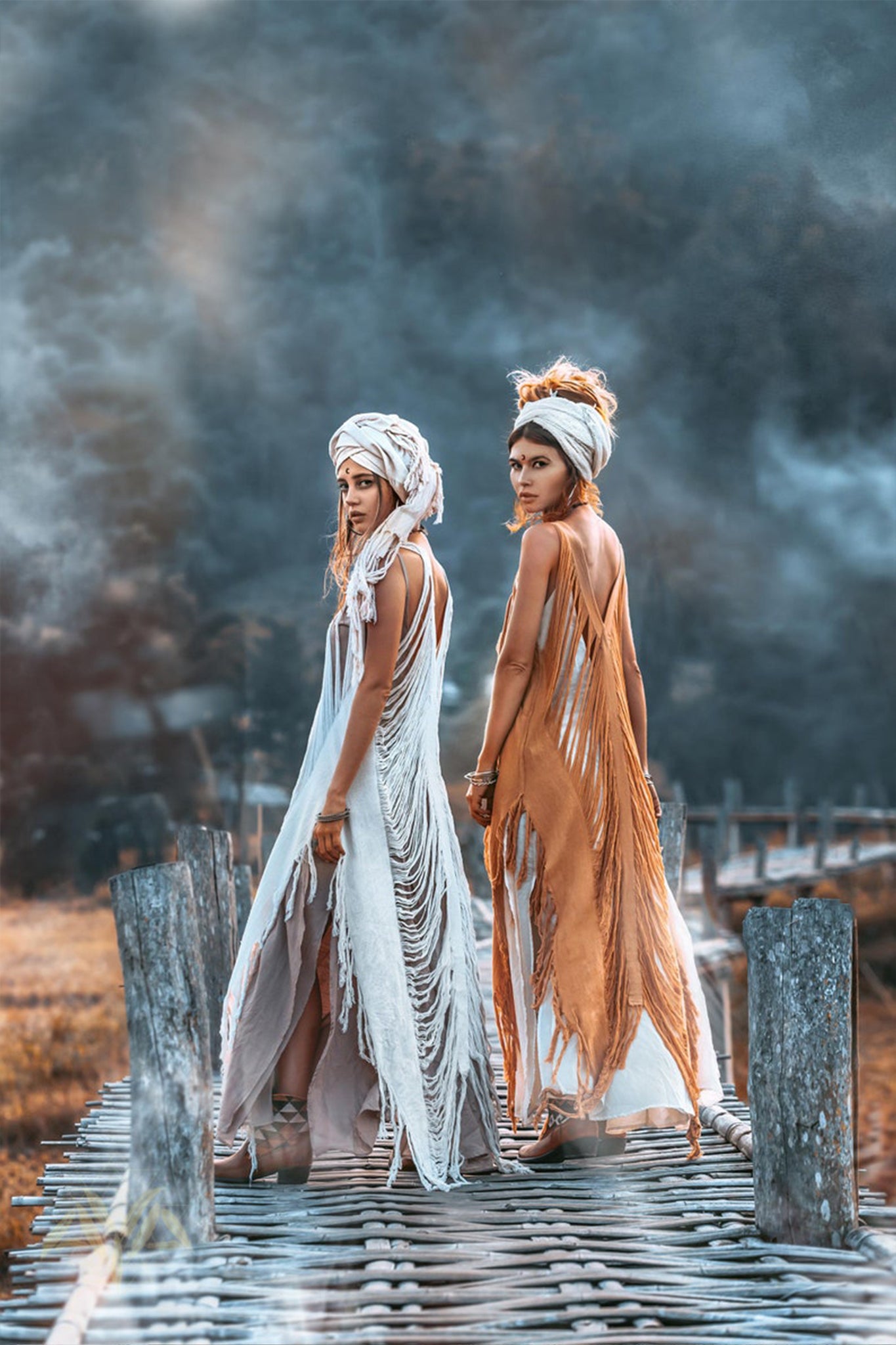 Two women pose on a wooden path, adorned in fringed, earth-toned See Through Dresses from AYA Sacred Wear's Long Maxi Dress collection. Each dress features exquisite handmade embroidery paired with elegant headwraps. They glance back at the camera amidst an atmospheric, misty backdrop that enhances the mystical ambiance.